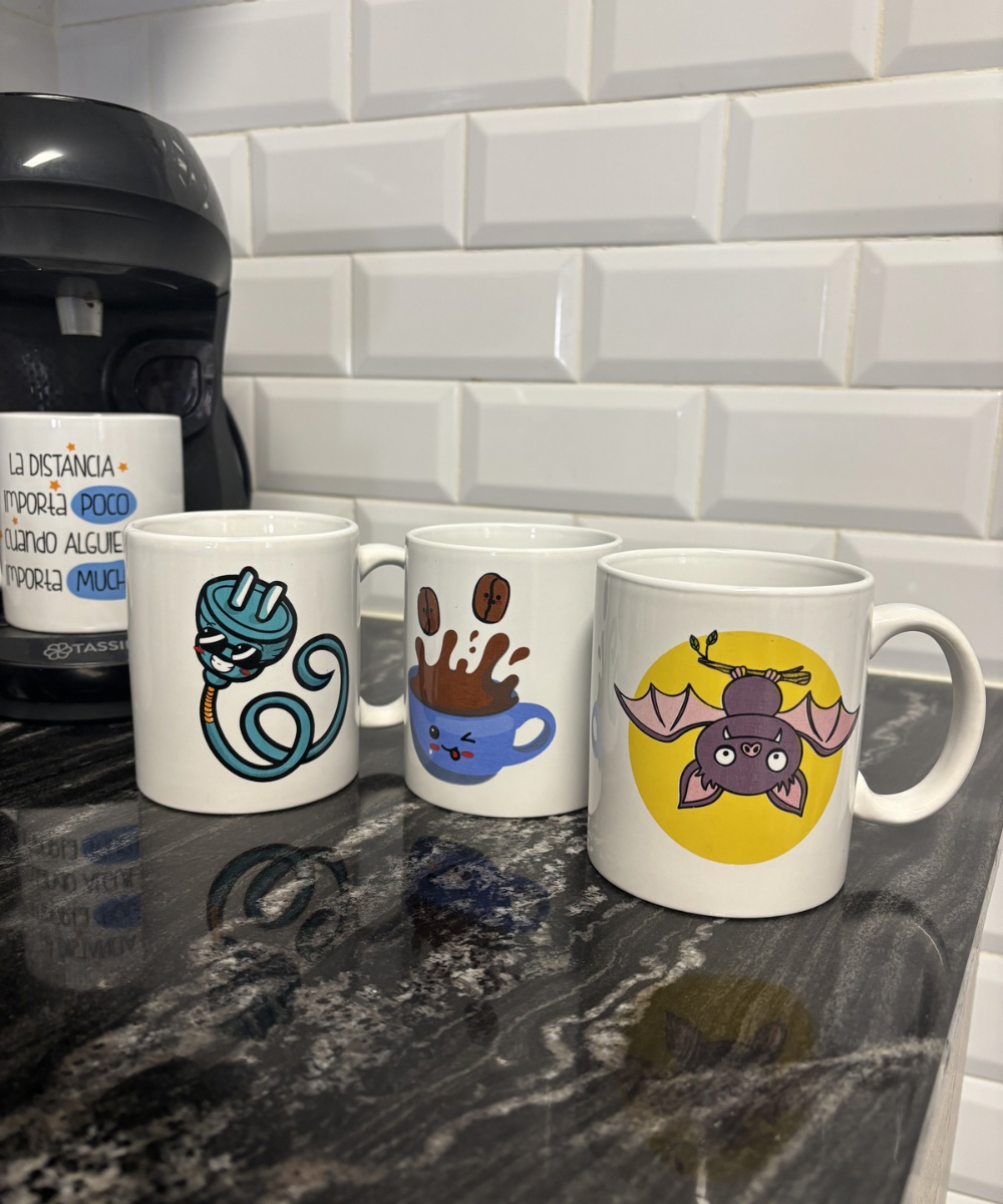 Cuatro tazas en una cocina, tres con dibujos divertidos y una colocada sobre la cafetera con una frase, en una composición doméstica.