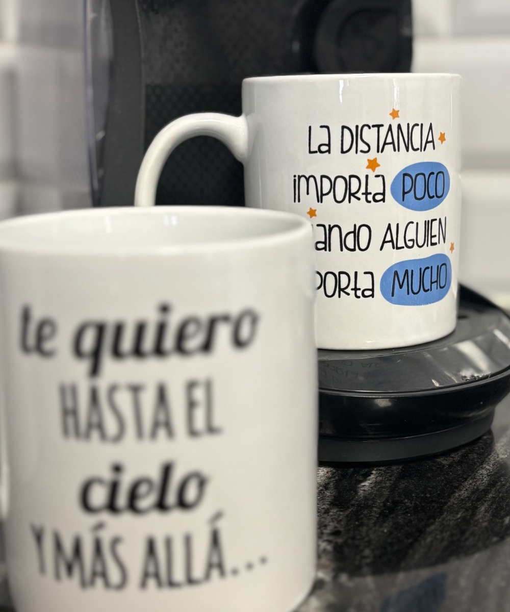 Dos tazas con frases, una en primer plano y otra ligeramente al fondo, colocadas en una cocina con una cafetera visible en el entorno.