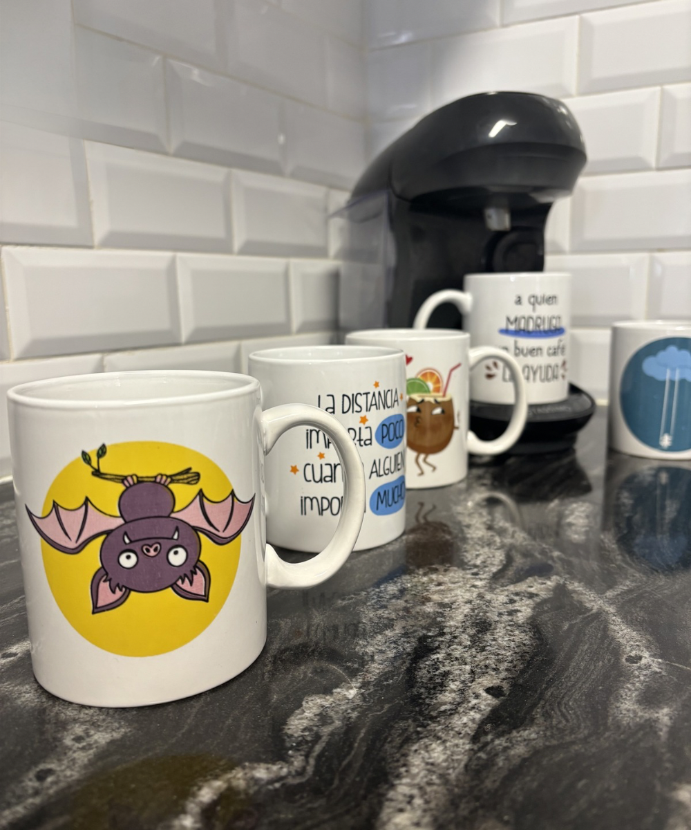 Cinco tazas divertidas con diferentes diseños, algunas con frases y otras con dibujos, colocadas en una cocina con una cafetera de fondo.